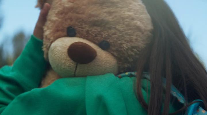 Face of brown teddy bear being held by a girl laying on shoulder of a green hoodie.  The girl has long dark straight hair.